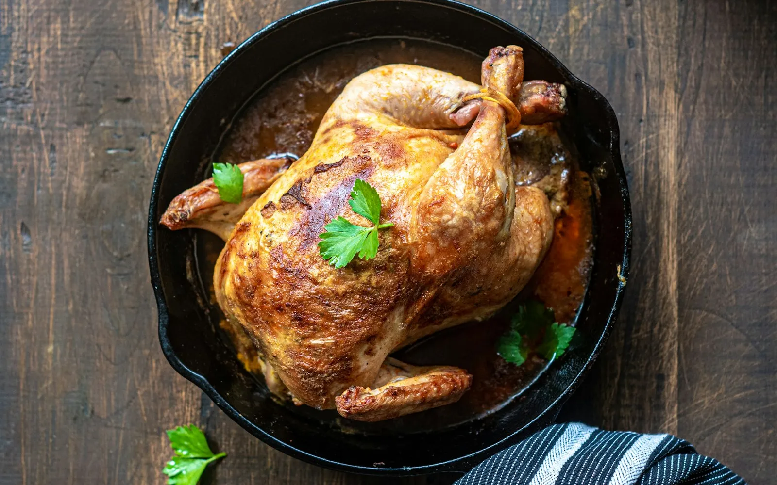 A golden roast chicken in a black cast-iron skillet on a dark wooden table