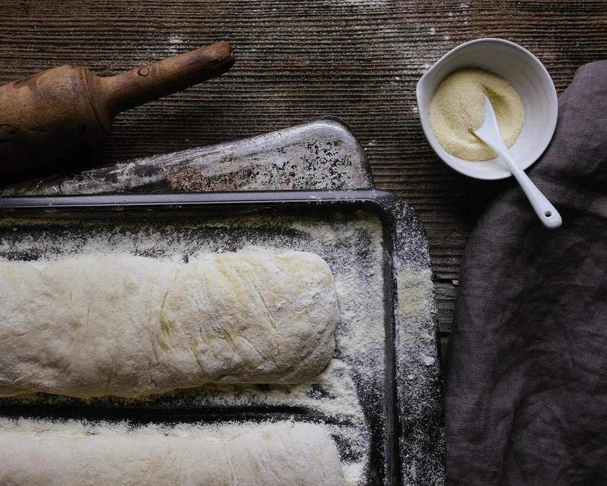 Shaped bread dough on a floured sheet pan next to a rolling pin and a bowl of semolina